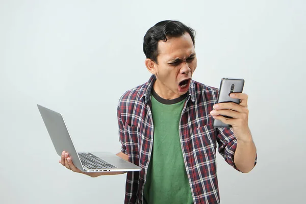 Angry mad asian indonesian man holding smartphone and laptop computer on isolated background