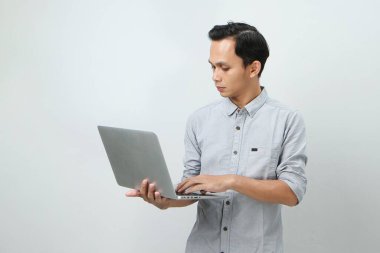 Asyalı endonezyalı genç adam elinde dizüstü bilgisayarla ciddi bir yüz ifadesiyle bir soru soruyor. Tecrit edilmiş arka planda kafa karıştırıcı bir fikir hakkında düşünceli.