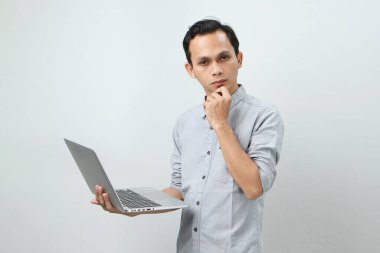 Asyalı endonezyalı genç adam elinde dizüstü bilgisayarla ciddi bir yüz ifadesiyle bir soru soruyor. Tecrit edilmiş arka planda kafa karıştırıcı bir fikir hakkında düşünceli.