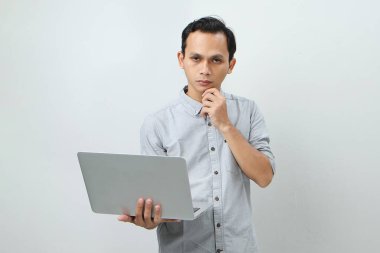 Asyalı endonezyalı genç adam elinde dizüstü bilgisayarla ciddi bir yüz ifadesiyle bir soru soruyor. Tecrit edilmiş arka planda kafa karıştırıcı bir fikir hakkında düşünceli.