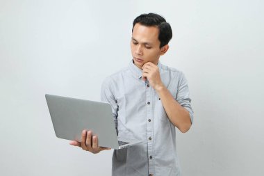 Asyalı endonezyalı genç adam elinde dizüstü bilgisayarla ciddi bir yüz ifadesiyle bir soru soruyor. Tecrit edilmiş arka planda kafa karıştırıcı bir fikir hakkında düşünceli.
