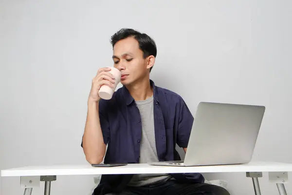 stock image asian man drinking from a cup at workplace while sitting in front of laptop computer. isolated background