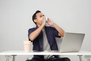 Asyalı adamın burnu akıyor, iş yerinde dizüstü bilgisayarın önünde otururken burnunu kağıt mendille siliyor. izole arkaplan