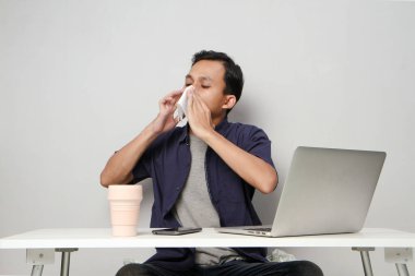 Asyalı adamın burnu akıyor, iş yerinde dizüstü bilgisayarın önünde otururken burnunu kağıt mendille siliyor. izole arkaplan