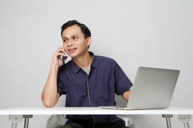 Neşeli mutlu Asyalı adam dizüstü bilgisayarın önünde otururken bir telefon görüşmesi yapıyor. izole edilmiş arkaplanda