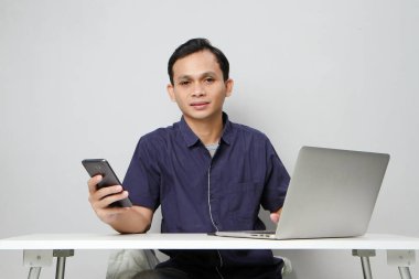 Dizüstü bilgisayarın önünde otururken cep telefonu tutan mutlu Asyalı adam. izole edilmiş arkaplanda