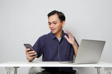 Dizüstü bilgisayarın önünde otururken cep telefonu tutan mutlu Asyalı adam. izole edilmiş arkaplanda