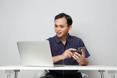 Dizüstü bilgisayarın önünde otururken cep telefonu tutan mutlu Asyalı adam. izole edilmiş arkaplanda