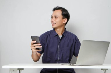 Dizüstü bilgisayarın önünde otururken cep telefonu tutan mutlu Asyalı adam. izole edilmiş arkaplanda