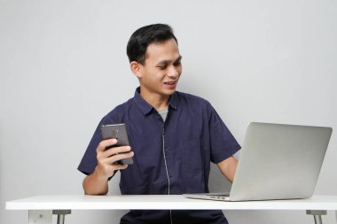 Dizüstü bilgisayarın önünde otururken cep telefonu tutan mutlu Asyalı adam. izole edilmiş arkaplanda