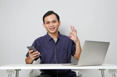 Cep telefonu tutan mutlu Asyalı adam dizüstü bilgisayarın önünde otururken işaret işareti yapıyor. izole edilmiş arkaplanda