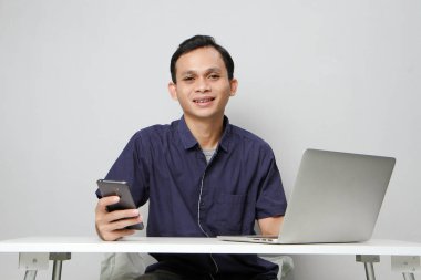 Dizüstü bilgisayarın önünde otururken cep telefonu tutan mutlu Asyalı adam. izole edilmiş arkaplanda