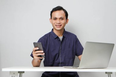 Dizüstü bilgisayarın önünde otururken cep telefonu tutan mutlu Asyalı adam. izole edilmiş arkaplanda