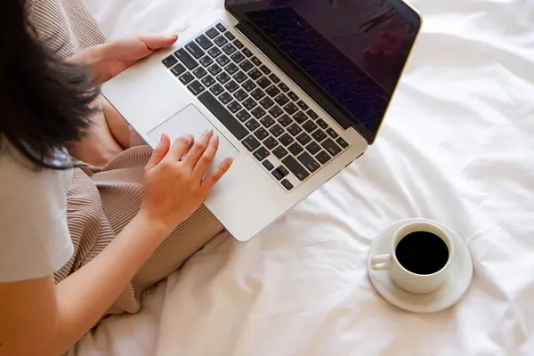 top view of Asian Woman sitting on the bed with cup of coffee and working on her laptop. with copy space