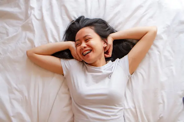 Top view of happy asian young woman lying in bed. Head shot portrait top view smiling beautiful woman lying in comfortable bed, looking at camera, young female wearing pajamas resting on soft pillows in bedroom, enjoying morning after awakenin