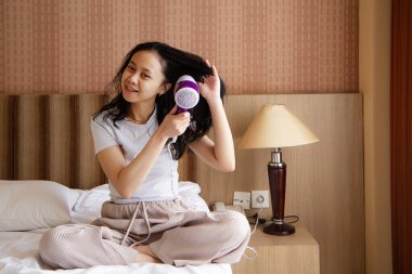 Yatakta oturan Asyalı kadın saç kurutuyor. Genç Asyalı kadın sabahları saç kurutma makinesi kullanıyor..