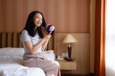 Yatakta oturan Asyalı kadın saç kurutuyor. Genç Asyalı kadın sabahları saç kurutma makinesi kullanıyor..