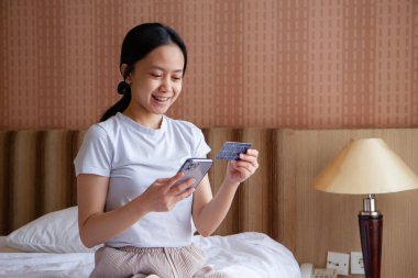 Asyalı kız evden online alışveriş yapıyor. Yatakta kredi kartı ve akıllı telefonuyla oturan genç bir kadın. Cep telefonu uygulamasında kadın siparişi, çevrimiçi ödeme