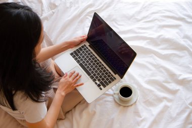 Asyalı kadının bir fincan kahveyle yatakta oturup dizüstü bilgisayarı üzerinde çalışmasının en üst görüntüsü. kopyalama alanı ile