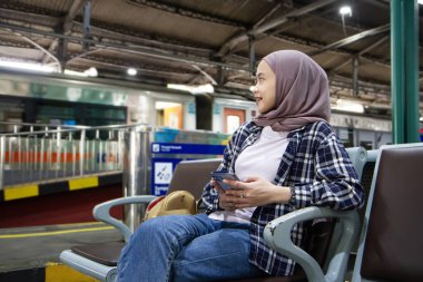 Mutlu Asyalı kadın cep telefonunu tutarken tren istasyonunun bekleme odasında oturuyor. seyahat kavramı