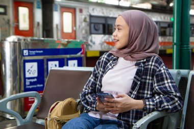 Mutlu Asyalı kadın cep telefonunu tutarken tren istasyonunun bekleme odasında oturuyor. seyahat kavramı