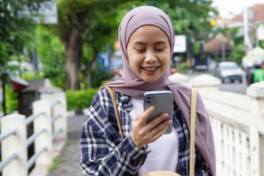 Mutlu Asyalı Müslüman kadın cep telefonuna bakarken sokakta yürüyor.