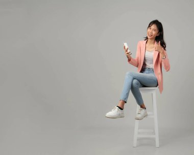 Mutlu, heyecanlı Asyalı bir kadının cep telefonuyla taburede oturması ve izole edilmiş arka planda el hareketi yapması.