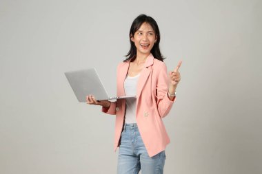 Mutlu Asyalı kadın resmi kıyafet giyiyor dizüstü bilgisayarı tutuyor ve arka planda parmak işareti yapıyor.