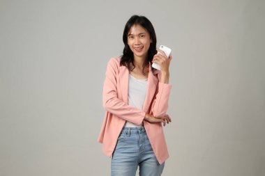 Mutlu Asyalı kadın resmi kıyafet giyiyor cep telefonunu kapalı bir arka planda tutuyor.