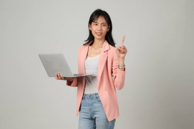 Mutlu Asyalı kadın resmi kıyafet giyiyor dizüstü bilgisayarı tutuyor ve arka planda parmak işareti yapıyor.