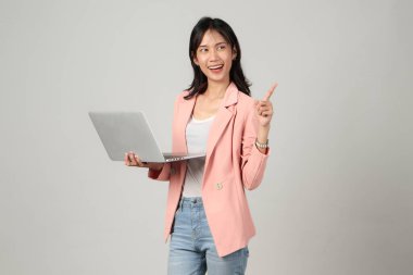 Mutlu Asyalı kadın resmi kıyafet giyiyor dizüstü bilgisayarı tutuyor ve arka planda parmak işareti yapıyor.