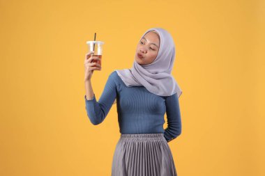 Mutlu Asyalı Müslüman kadın izole edilmiş sarı arka planda plastik bardakta buzlu çay tutuyor ve içiyor.