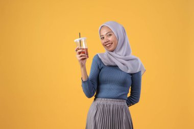 Mutlu Asyalı Müslüman kadın izole edilmiş sarı arka planda plastik bardakta buzlu çay tutuyor ve içiyor.