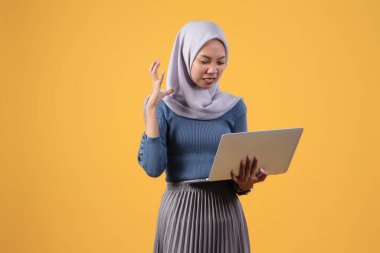 Asyalı Endonezyalı Müslüman kadın izole edilmiş sarı arka planda kızgın yüz ifadesi olan dizüstü bilgisayarı tutuyor.
