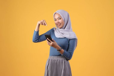 Mutlu Asyalı Müslüman kadın, izole edilmiş sarı arka planda işaret eden akıllı bir telefon tutuyor.