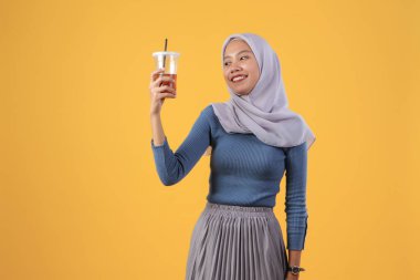 Mutlu Asyalı Müslüman kadın izole edilmiş sarı arka planda plastik bardakta buzlu çay tutuyor ve içiyor.