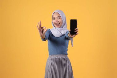 Mutlu Asyalı Müslüman kadın akıllı telefonu tutuyor ve akıllı telefon boş ekranını gösteriyor ve izole edilmiş sarı arka planda parmak hareketi yapıyor.