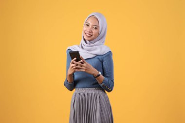 Mutlu Asyalı Müslüman kadın izole edilmiş sarı arka planda akıllı telefonu tutuyor.