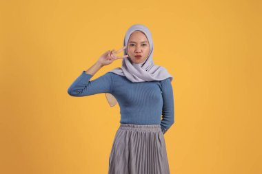 Mutlu Asyalı Müslüman kadın izole edilmiş sarı arka planda zafer işareti yapıyor.