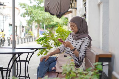 Asyalı Müslüman kadınlar, Kota Lama ya da eski şehir Semarang, Endonezya 'da açık kahve dükkanındaki bankta otururken akıllı telefonuna bakıyorlar.