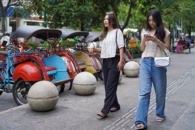 İki Asyalı kadın ellerinde cep telefonuyla sokakta yürüyor. Malioboro Caddesi, Yogyakarta, Endonezya