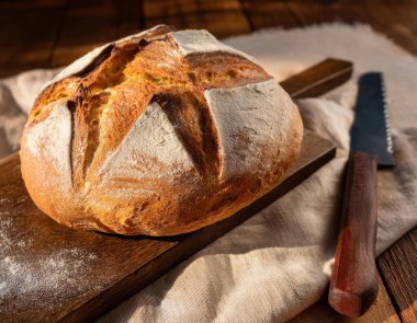 Ekmek bıçağıyla ahşap bir kesme tahtasına yerleştirilmiş, altından kabuklu bir ekmek somunu, dağınık un ve sıcak, doğal bir arka plan üzerine kurulmuş keten bir bez parçası.