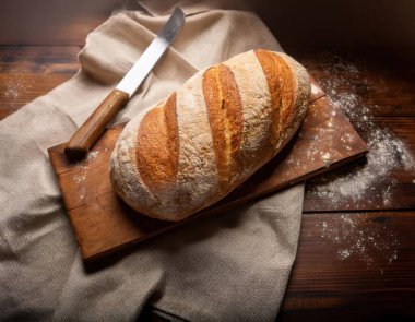 Ekmek bıçağıyla ahşap bir kesme tahtasına yerleştirilmiş, altından kabuklu bir ekmek somunu, dağınık un ve sıcak, doğal bir arka plan üzerine kurulmuş keten bir bez parçası.