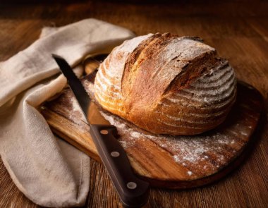 Ekmek bıçağıyla ahşap bir kesme tahtasına yerleştirilmiş, altından kabuklu bir ekmek somunu, dağınık un ve sıcak, doğal bir arka plan üzerine kurulmuş keten bir bez parçası.