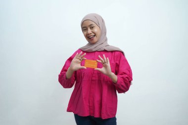 Bej tesettür ve pembe bluz giyen genç Asyalı Müslüman kadın gülümsüyor ve izole edilmiş arka planda turuncu bir kredi kartı sunuyor. Pazarlama, bankacılık ve dijital finans temaları için ideal.