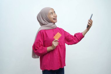 Neşeli genç Müslüman kadın bej tesettür ve parlak pembe bluz giyerek elinde akıllı telefon ve izole edilmiş arka planda turuncu bir kredi kartı tutarken gülümsüyor..