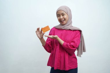 Bej tesettür ve pembe bluz giyen genç Asyalı Müslüman kadın gülümsüyor ve izole edilmiş arka planda turuncu bir kredi kartı sunuyor. Pazarlama, bankacılık ve dijital finans temaları için ideal.