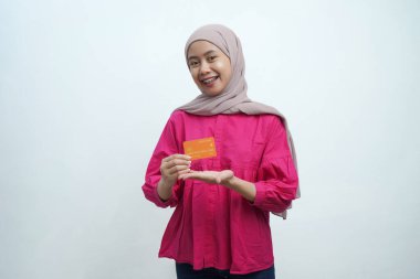 Bej tesettür ve pembe bluz giyen genç Asyalı Müslüman kadın gülümsüyor ve izole edilmiş arka planda turuncu bir kredi kartı sunuyor. Pazarlama, bankacılık ve dijital finans temaları için ideal.