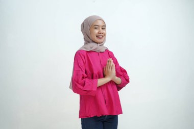 Genç Asyalı Müslüman kadın açık kahverengi bir başörtüsü ve parlak pembe bir bluz giyerek, her iki avuçla birlikte izole bir arka planda geleneksel bir selamlama hareketi yaparken sıcacık bir şekilde gülümsüyor..