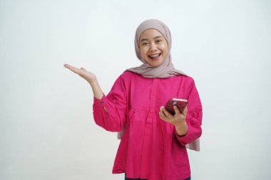 Pembe bluz ve açık kahverengi tesettürlü neşeli genç Müslüman kadın bir elinde akıllı bir telefon tutarken gülümsüyor diğer elinde ise sanki bir şey sunuyor ya da sergiliyormuş gibi el kol hareketi yapıyor. izole edilmiş arkaplanda